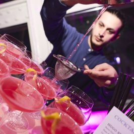 Bar tender pouring a cocktail from a height in front of table of pink cocktails