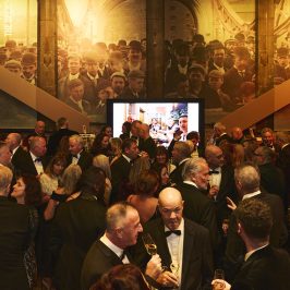 Crowd of people in smart dress in the North Tower Level Four at Tower Bridge with a screen showing personal images